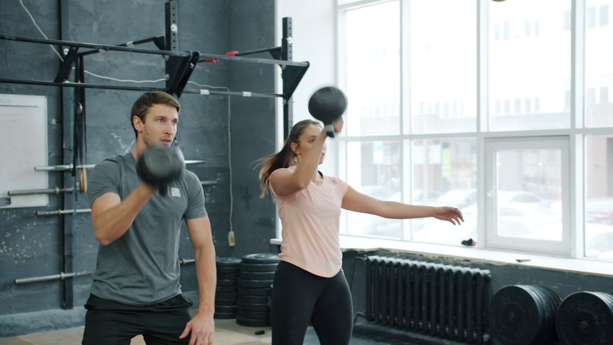 Girl and guy sports partners are squatting and lifting heavy kettlebells working out in gym together focused on exercise. People are wearing trendy sportswear.
